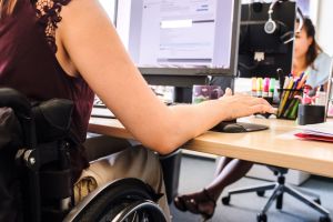 Bild von einer Frau die am Schreibtisch arbeitet und dabei im Rollstuhl sitzt.